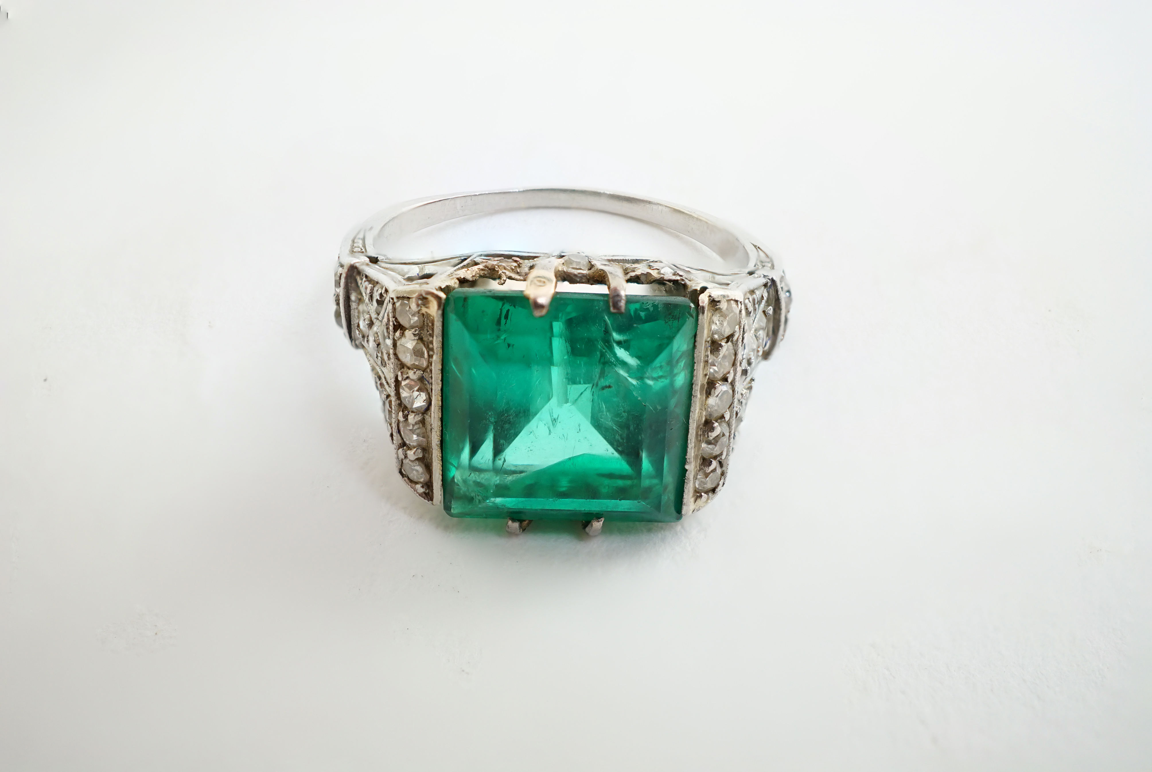 A 1920's/1930's platinum and square cut emerald set dress ring, with pierced and millegrain diamond cluster set shoulders